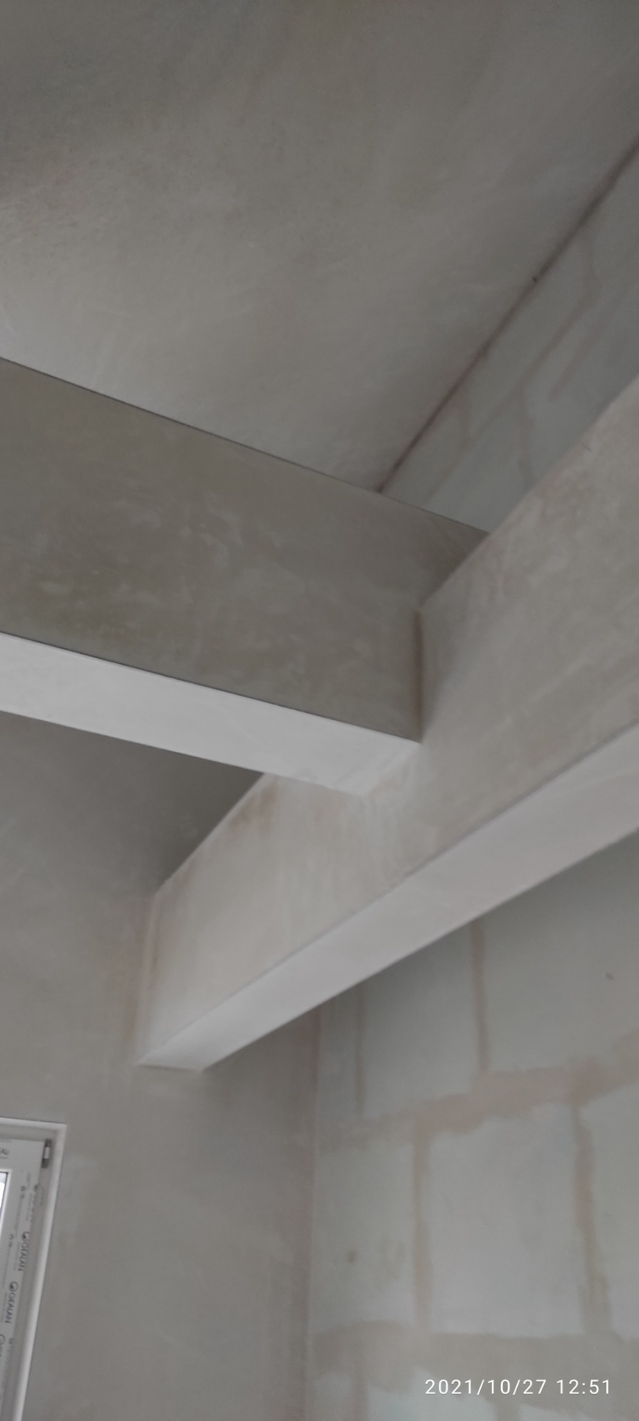 Szare ściany i sufit po nałożeniu tynku maszynowego, widoczne elementy konstrukcyjne pod sufitem, ujęcie z dołu, w rogu okno.