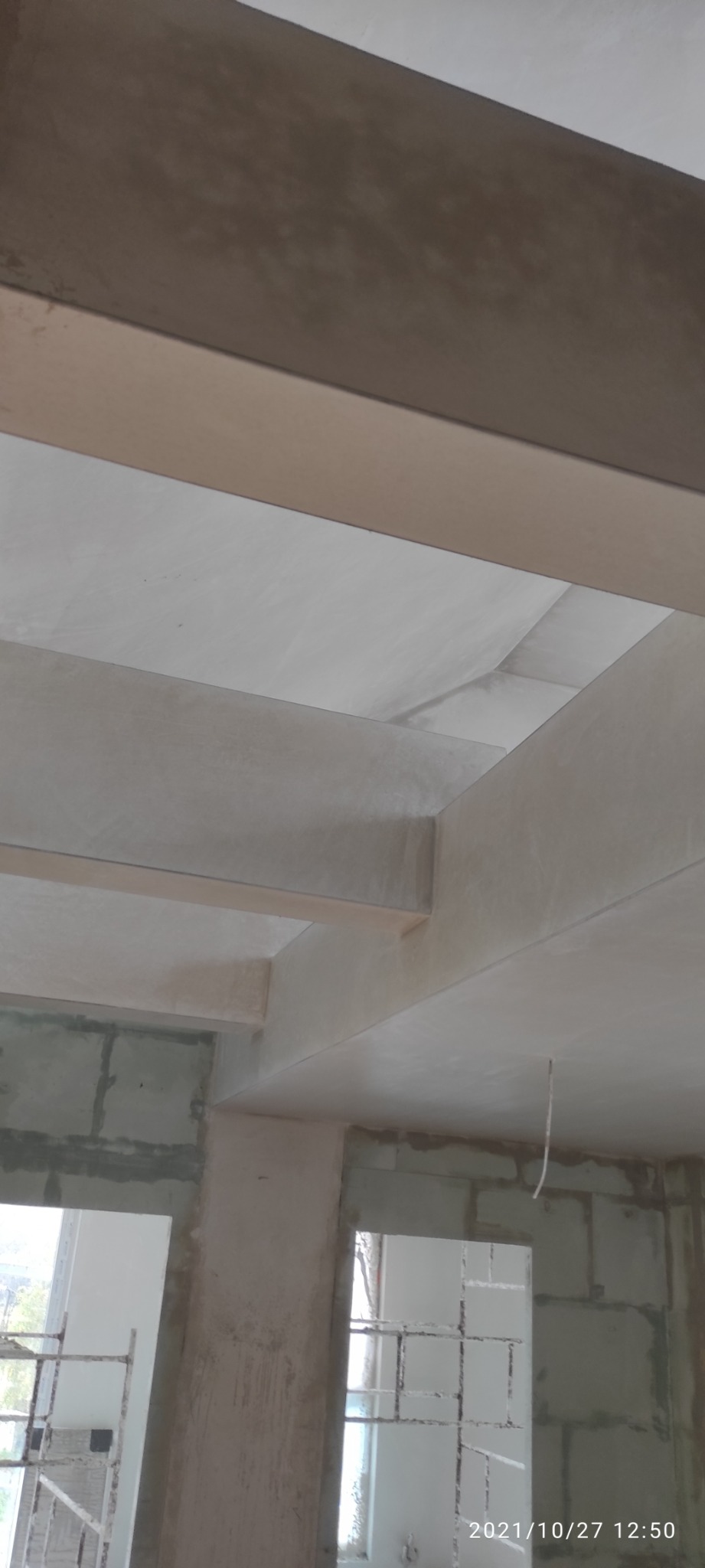 Surowy stan wnętrza z widocznymi elementami konstrukcyjnymi stropu i ścian po nałożeniu warstwy tynku, z wiszącym przewodem elektrycznym i blokami betonowymi w tle.