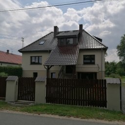 Am Bud Adam Kozak - Dwukondygnacyjny dom jednorodzinny z beżową elewacją, ciemną dachówką i balkonem z ozdobną balustradą, widziany zza drewnianego ogrodzenia z betonowymi słupkami.