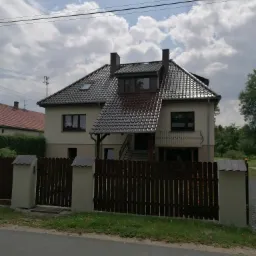 Dwukondygnacyjny dom jednorodzinny z beżową elewacją, ciemną dachówką i balkonem z ozdobną balustradą, widziany zza drewnianego ogrodzenia z betonowymi słupkami.