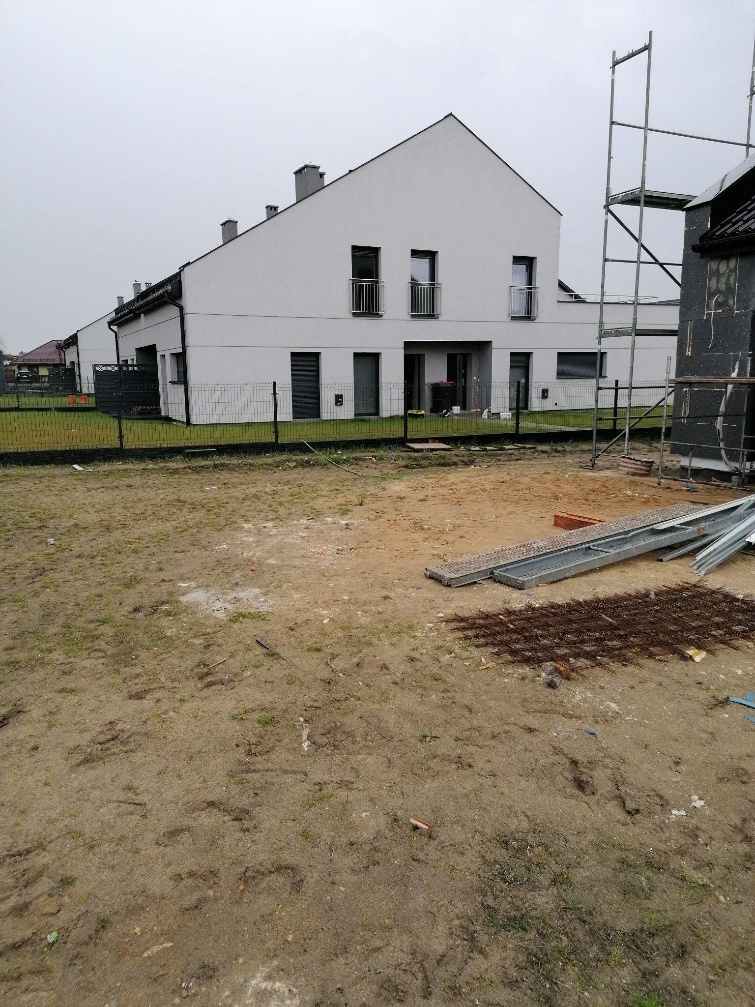 Nowoczesny dom w zabudowie szeregowej z białą elewacją, widoczne rusztowanie i materiały budowlane na niezagospodarowanym terenie przed budynkiem.