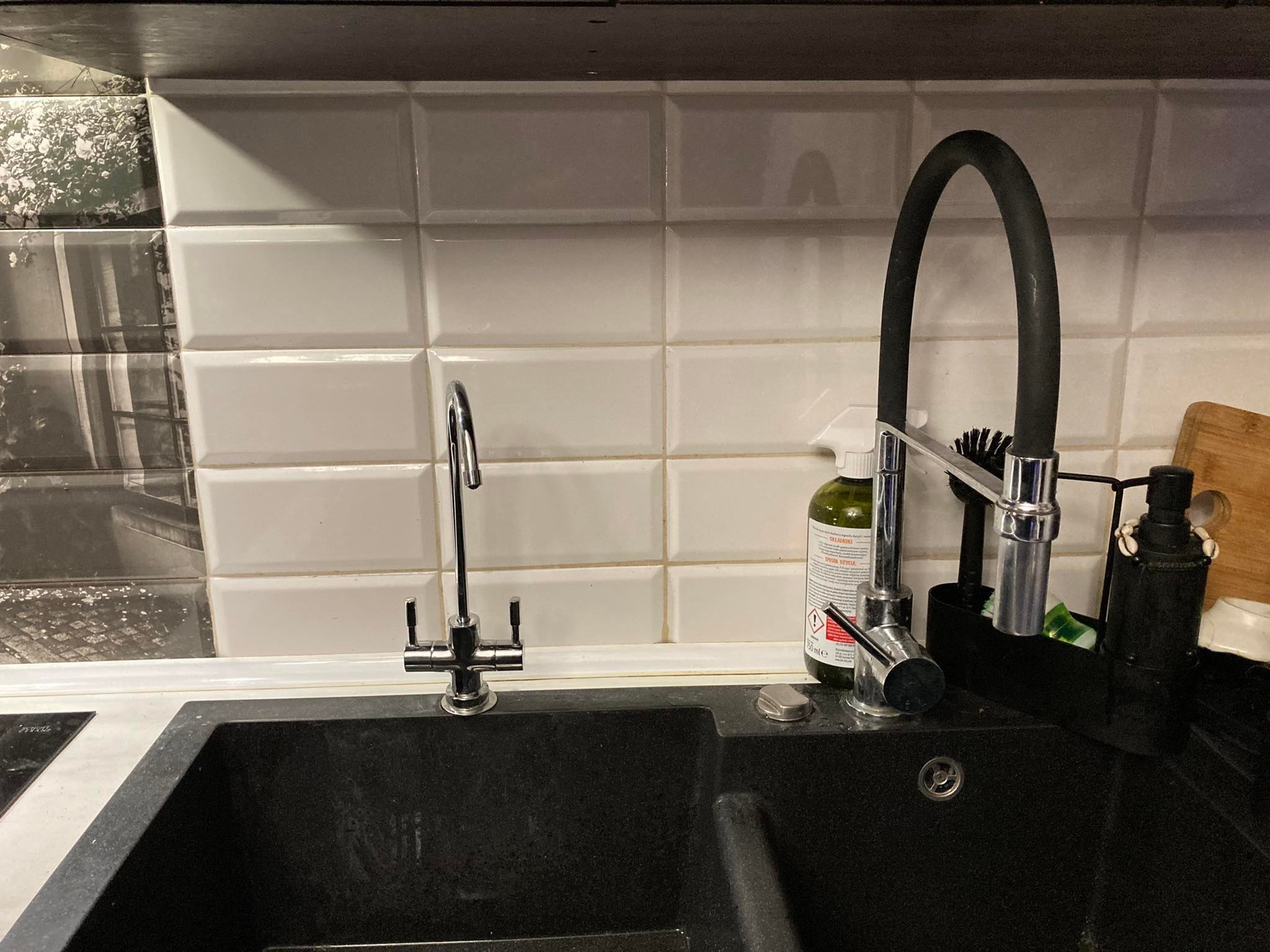 Czarny, dwukomorowy zlew granitowy z dwoma bateriami, dozownikiem na płyn i butelką detergentu na tle białych płytek z dekoracyjnym pasem.