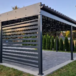 Nowoczesna, ciemna altana z metalu z poziomymi panelami przesłaniającymi, na kostce brukowej, otoczona zielenią i tujami.