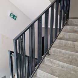 Mariusz Cierlik - Metalowa balustrada schodowa z pionowymi prętami o prostokątnym przekroju, zamontowana na schodach wyłożonych płytkami imitującymi kamień, z widocznym znakiem ewakuacyjnym na ścianie.