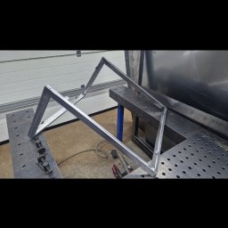 Mariusz Cierlik - Element konstrukcji metalowej w trakcie spawania, widoczne narzędzia i stół spawalniczy, surowe wykończenie.