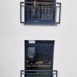Mariusz Cierlik - Elewacja budynku z dwoma oknami zabezpieczonymi prostymi, ciemnymi balustradami balkonowymi, widoczne odbicie otoczenia w szybach okiennych.