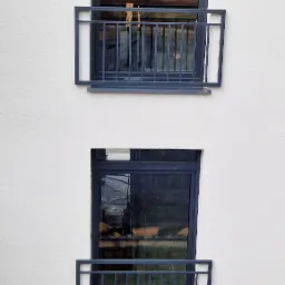 Elewacja budynku z dwoma oknami zabezpieczonymi prostymi, ciemnymi balustradami balkonowymi, widoczne odbicie otoczenia w szybach okiennych.