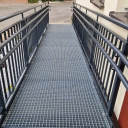 Mariusz Cierlik - Metalowy podjazd dla wózków inwalidzkich z balustradami w Trójcy, z kratową powierzchnią, prowadzący do budynku o elewacji w kolorze beżowo-czerwonym.