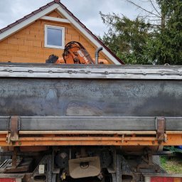 Mariusz Cierlik - Widok z tyłu pomarańczowej ciężarówki z zamontowaną metalową skrzynią ładunkową po spawaniu, z widocznymi śladami obróbki termicznej metalu i spoinami, na tle domu z cegły i drzew