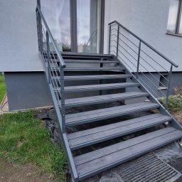 Mariusz Cierlik - Zewnętrzne schody metalowe z balustradą, prowadzące do wejścia domu. Szare stopnie z desek kompozytowych, minimalistyczna balustrada z poziomych prętów. Widok z dołu, zieleń w tle.
