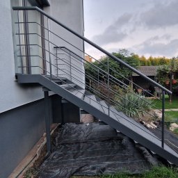 Mariusz Cierlik - Zewnętrzne schody metalowe z prostą balustradą, prowadzące do drzwi na piętrze. Widok z dołu, z ogrodem w tle. Minimalistyczny design, szara kolorystyka.