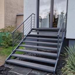 Mariusz Cierlik - Zewnętrzne schody metalowe z szarą balustradą, prowadzące do drzwi wejściowych budynku. Widoczne rośliny w otoczeniu schodów i fragment elewacji budynku.