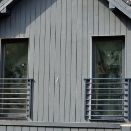 Mariusz Cierlik - Szara elewacja domu z dwoma oknami zabezpieczonymi poziomymi, metalowymi balustradami. Widok na fragment dachu i niebo.