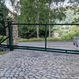 Mariusz Cierlik - Otwarta, zielona brama wjazdowa z kamiennymi słupkami, prowadząca na posesję z brukowaną nawierzchnią. W tle zieleń drzew i zabudowania.