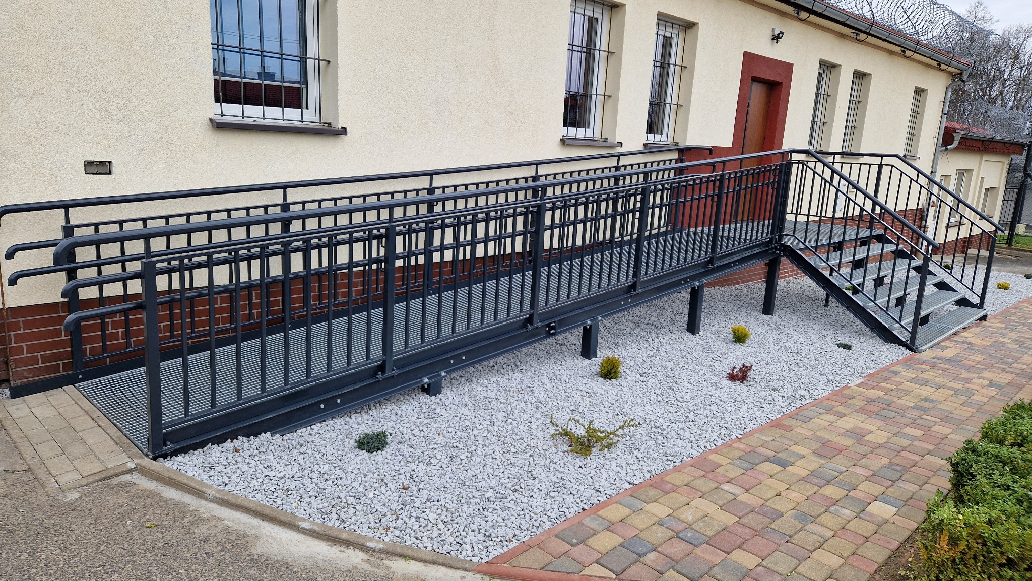 Zewnętrzna rampa dla niepełnosprawnych z metalową konstrukcją i balustradami, prowadząca do schodów, otoczona białym żwirem i kostką brukową.