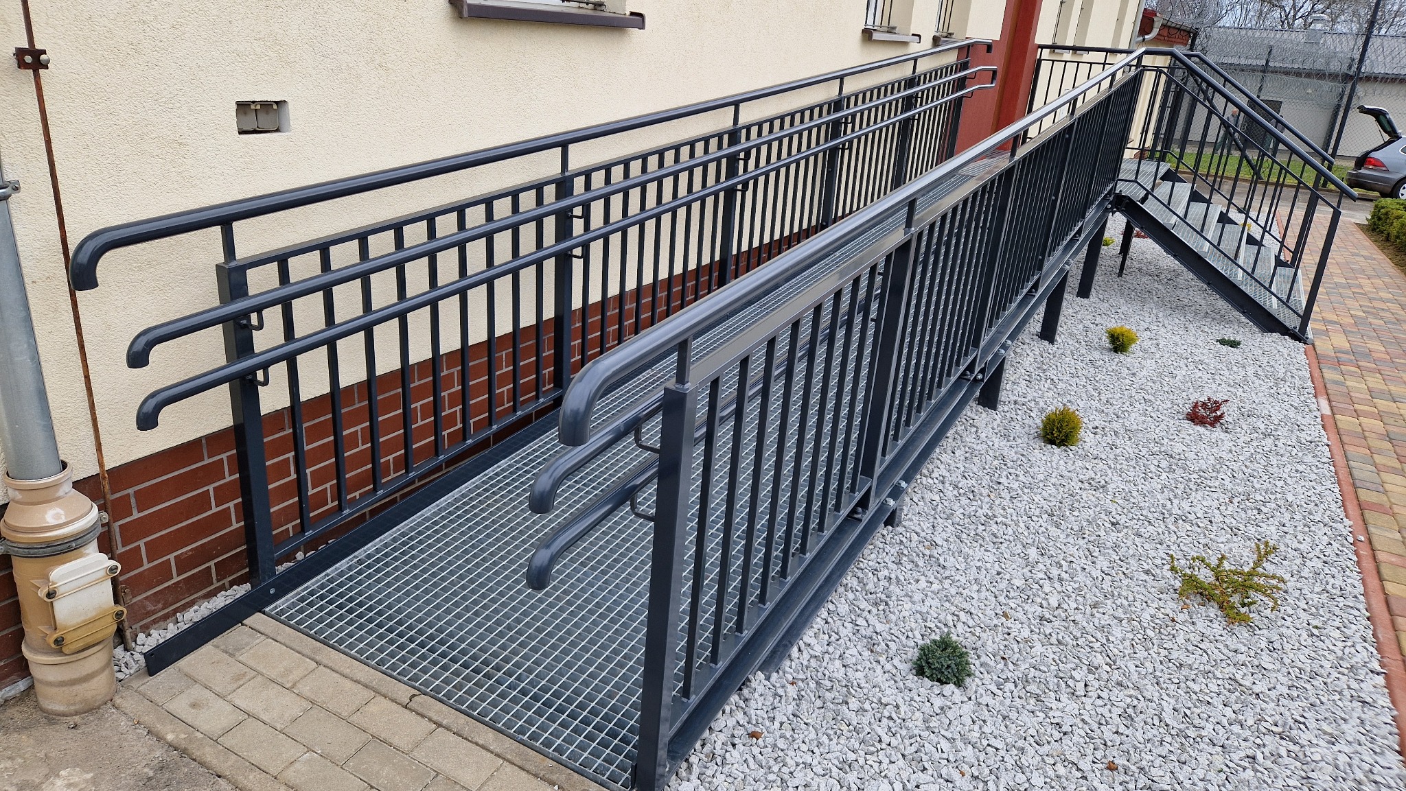 Metalowa rampa dla niepełnosprawnych z poręczami i kratą pomostową, prowadząca obok schodów na tle budynku z ceglaną wstawką i ozdobnym żwirowym podłożem.