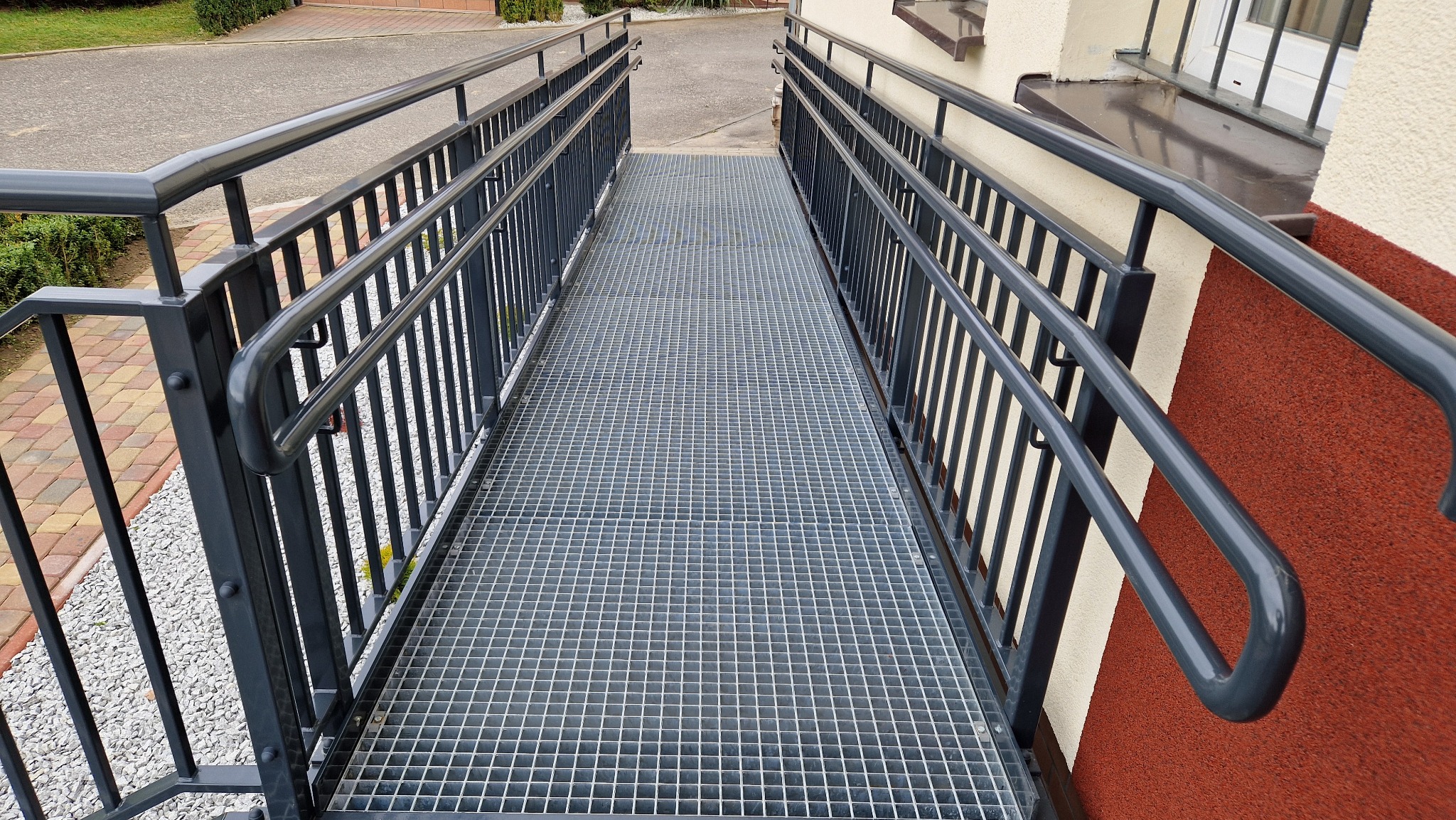 Metalowy podjazd dla wózków inwalidzkich z balustradami w Trójcy, z kratową powierzchnią, prowadzący do budynku o elewacji w kolorze beżowo-czerwonym.