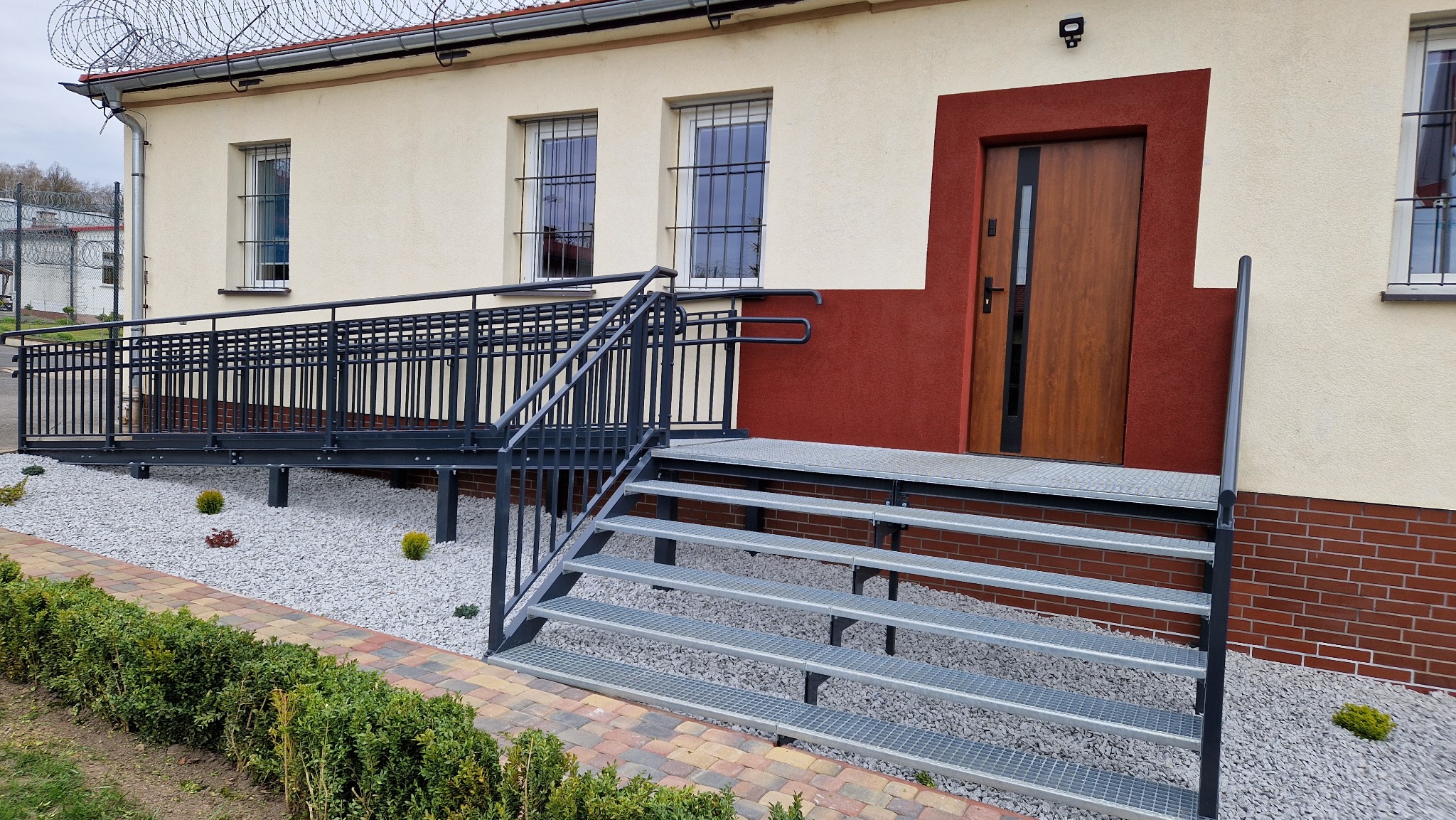 Zewnętrzne metalowe schody z kraty pomostowej i podjazdem dla niepełnosprawnych przed budynkiem z oknami zabezpieczonymi kratami.