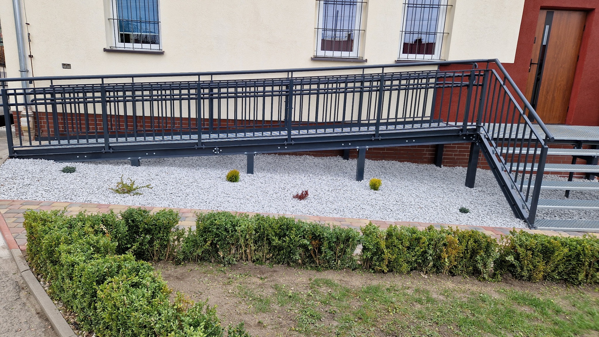 Zewnętrzna rampa z ciemnego metalu z balustradami, prowadząca do drzwi budynku, otoczona białym żwirem i niskimi krzewami.