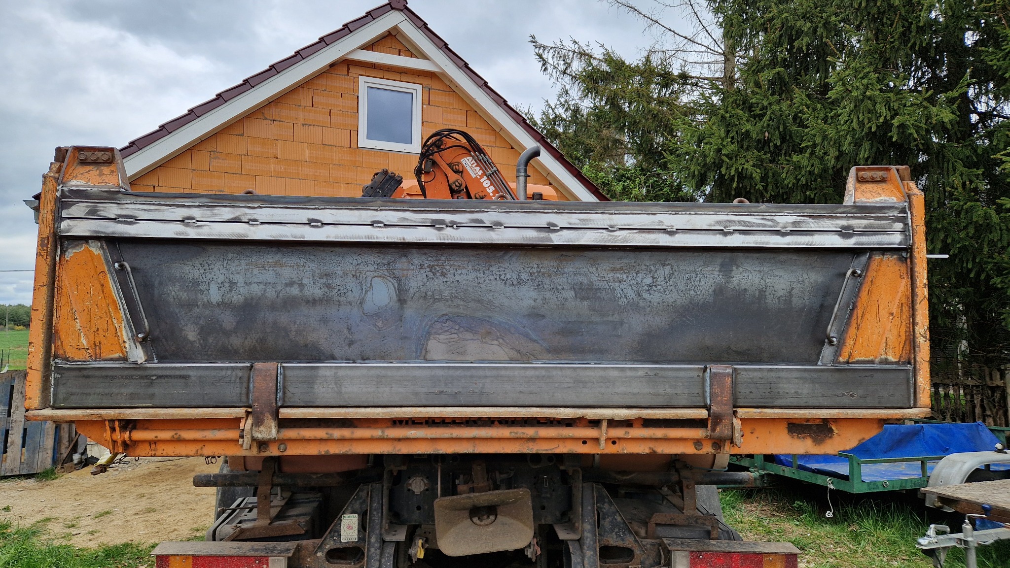 Widok z tyłu pomarańczowej ciężarówki z zamontowaną metalową skrzynią ładunkową po spawaniu, z widocznymi śladami obróbki termicznej metalu i spoinami, na tle domu z cegły i drzew