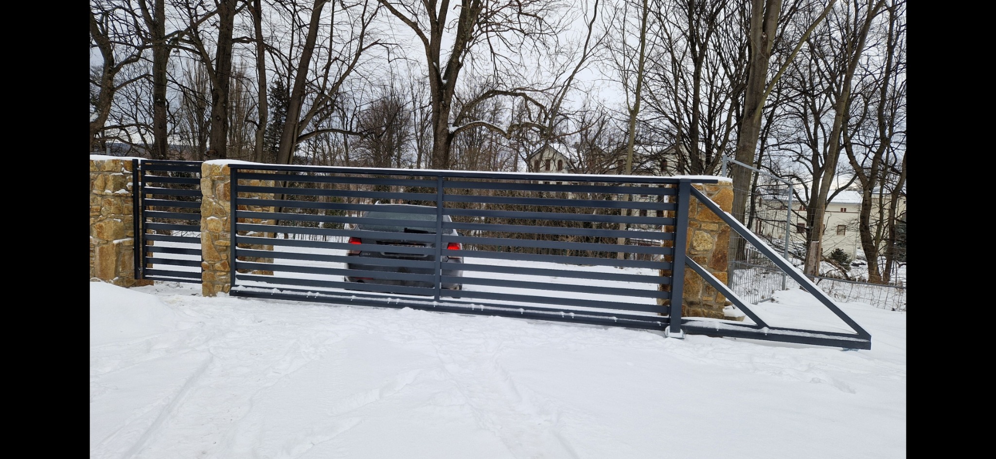 Nowoczesne ogrodzenie posesji z poziomych, grafitowych profili metalowych, osadzone na kamiennych słupkach, częściowo zasypane śniegiem, z widocznym samochodem w tle.