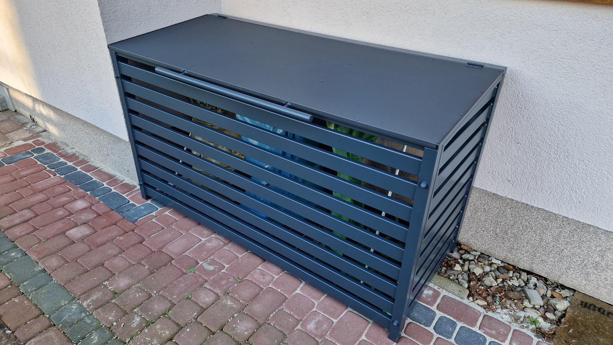 Metalowa obudowa na pojemniki na śmieci z poziomymi szczeblami, stojąca na kostce brukowej przy ścianie budynku.