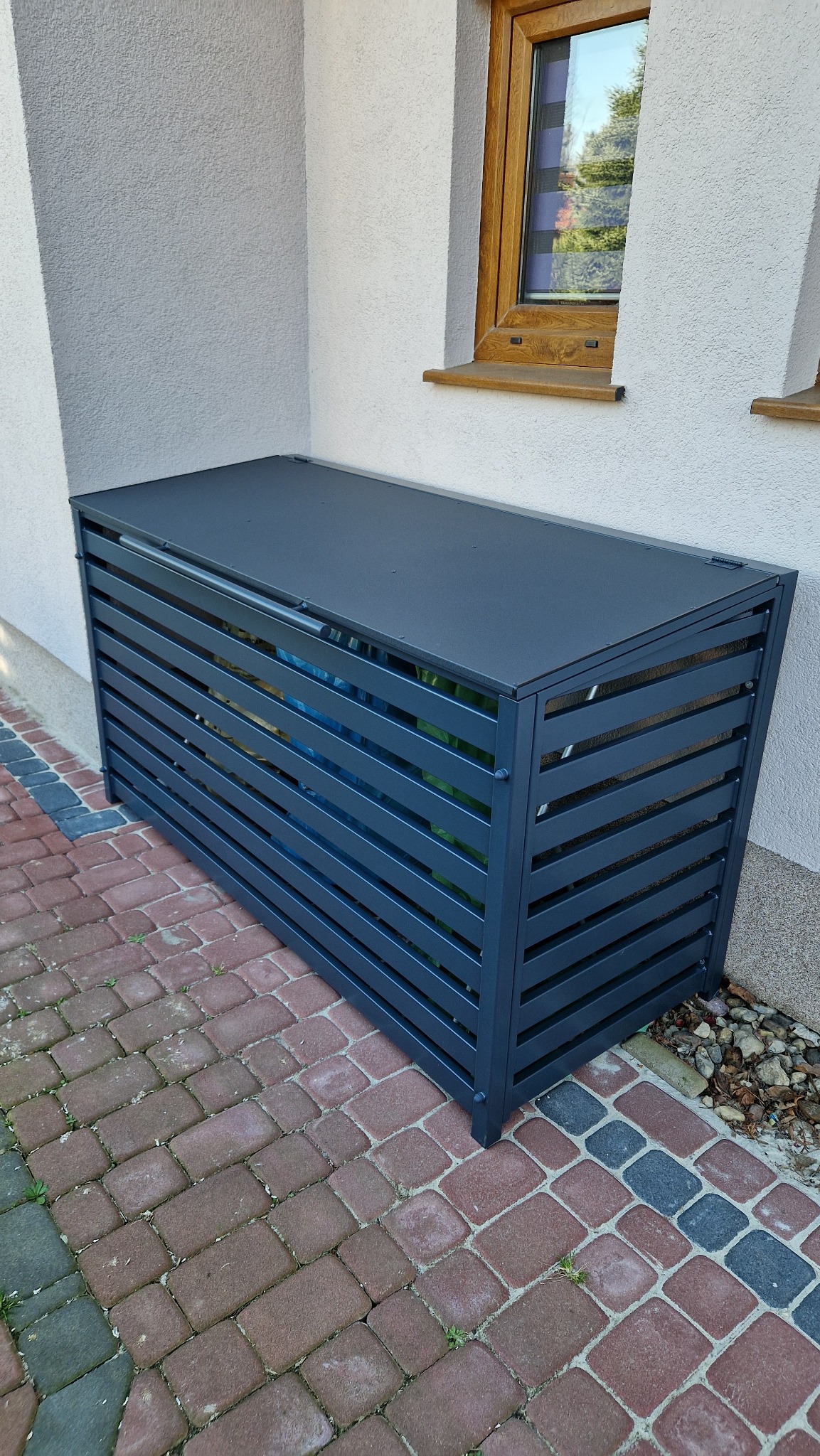 Metalowa obudowa na śmietnik z poziomymi szczeblami, stojąca na kostce brukowej przy ścianie domu z oknem.