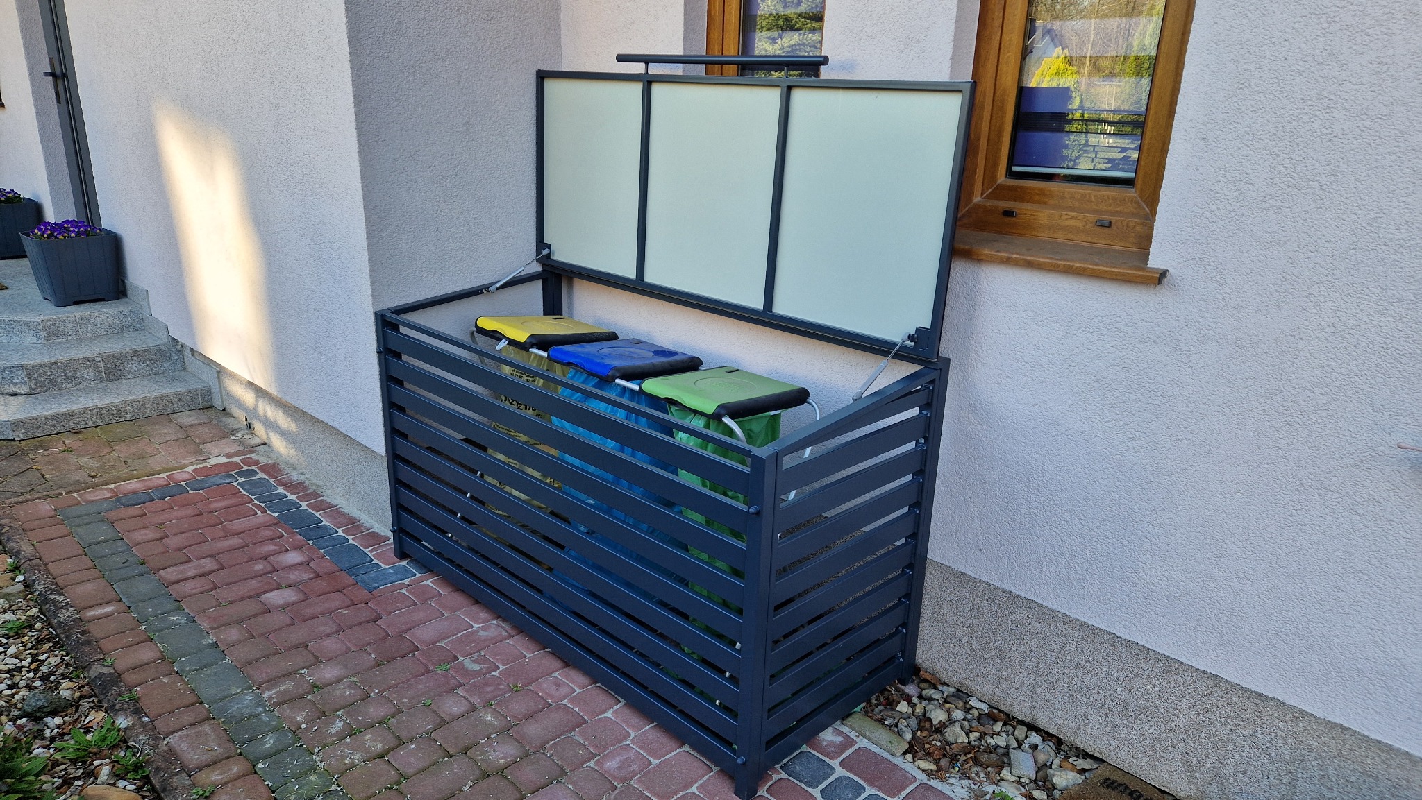 Zewnętrzna obudowa na pojemniki do segregacji śmieci, wykonana z szarej metalowej konstrukcji z poziomymi szczebelkami i uchylną klapą z białymi panelami, stojąca na kostce brukowej przy ścianie domu.