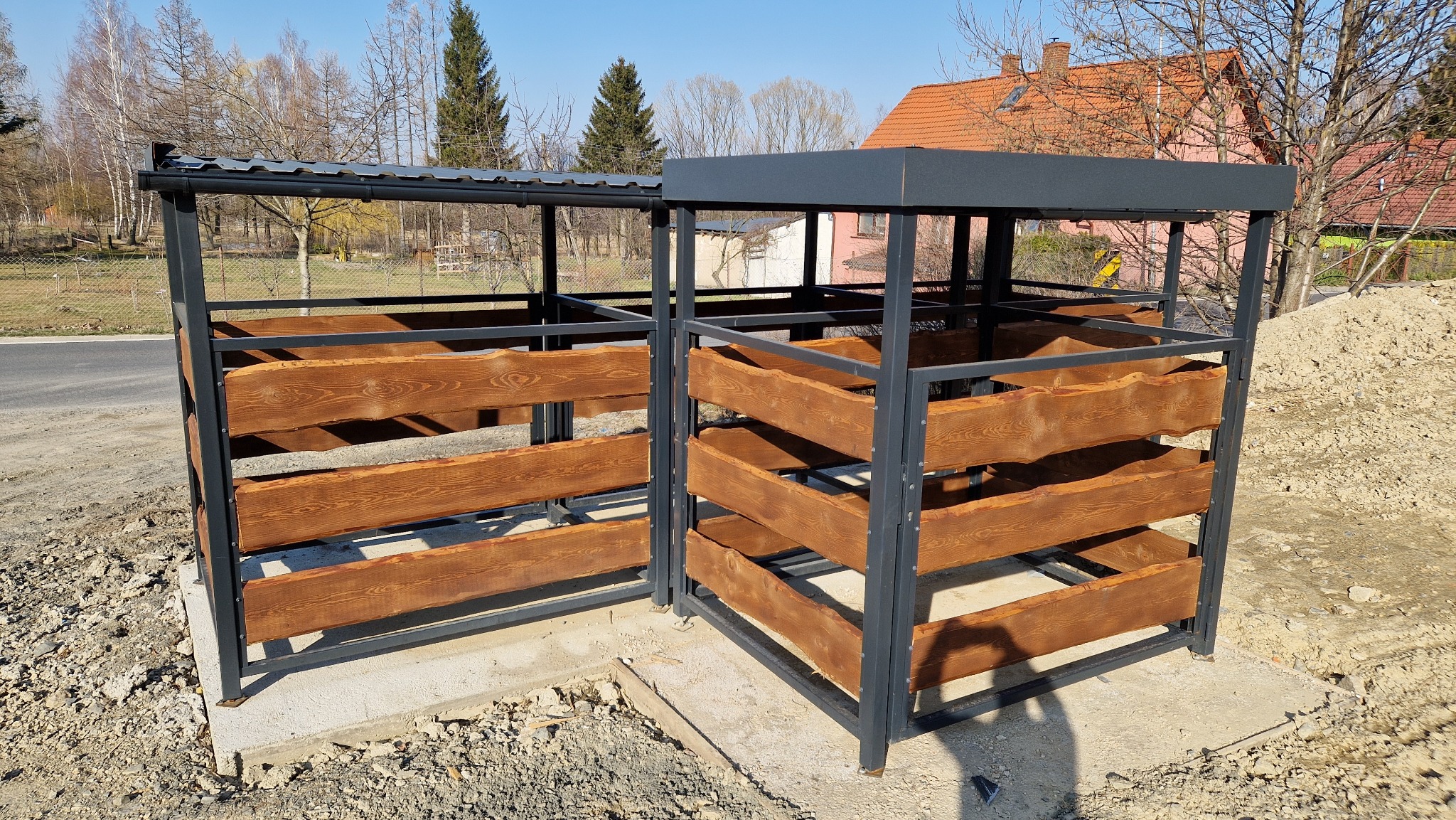 Dwa zadaszone pojemniki na śmieci z metalową ramą i drewnianymi deskami, ustawione na betonowej podstawie, z widocznym placem budowy w tle.