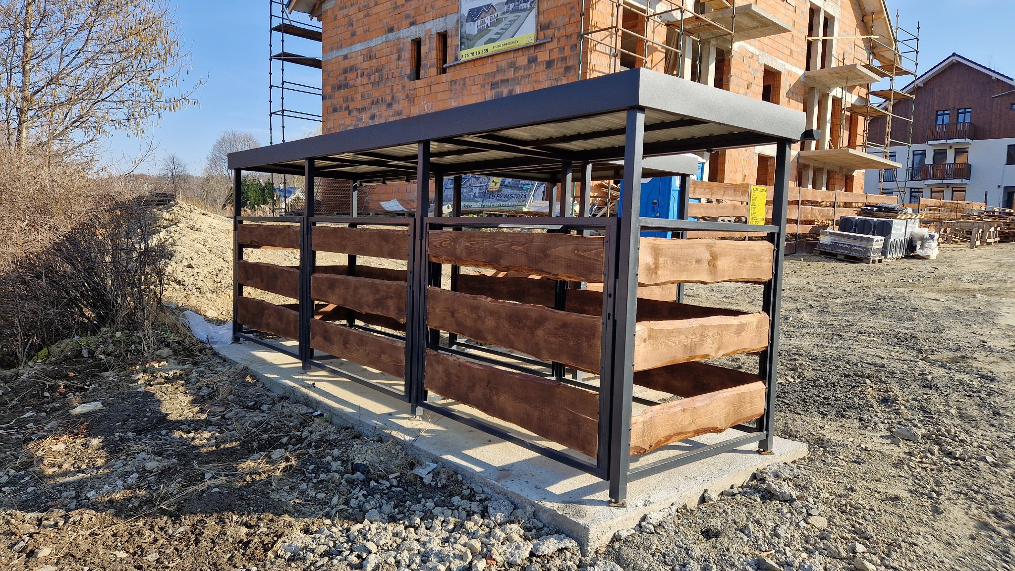 Metalowa konstrukcja z drewnianymi wypełnieniami, prawdopodobnie na pojemniki na odpady, na tle placu budowy z ceglanym budynkiem w stanie surowym i stertami materiałów.