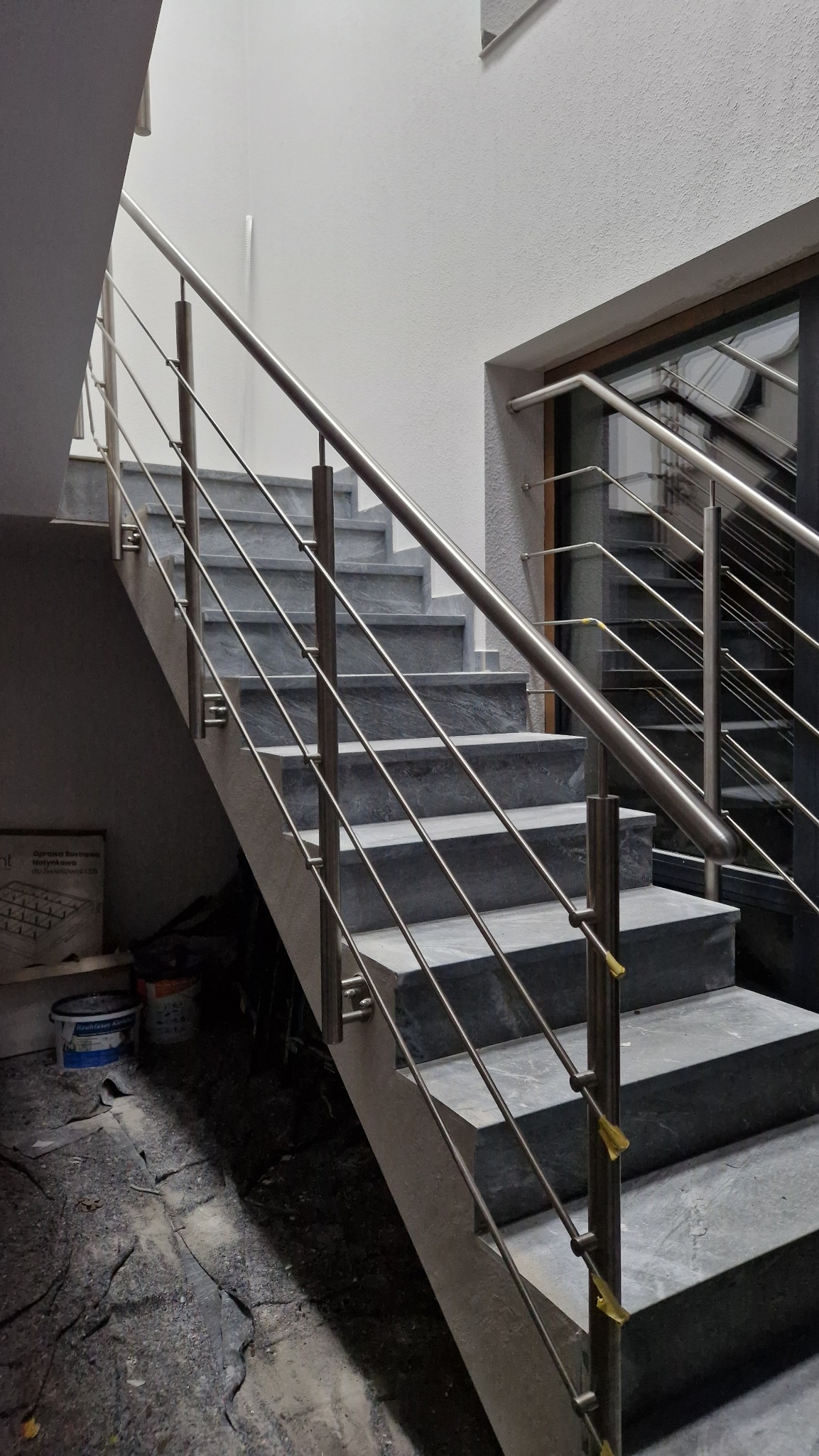 Szare schody z nierdzewną balustradą, widok z dołu, na podłodze widoczne ślady prac wykończeniowych, oklejone taśmą zabezpieczającą