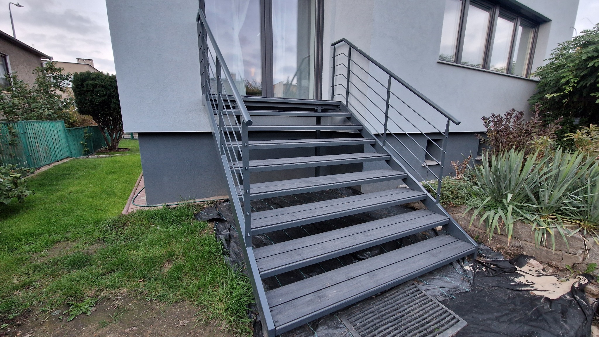 Zewnętrzne schody metalowe z balustradą, prowadzące do wejścia domu. Szare stopnie z desek kompozytowych, minimalistyczna balustrada z poziomych prętów. Widok z dołu, zieleń w tle.