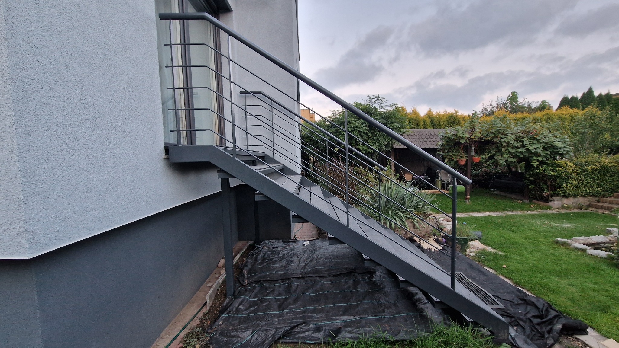 Zewnętrzne schody metalowe z prostą balustradą, prowadzące do drzwi na piętrze. Widok z dołu, z ogrodem w tle. Minimalistyczny design, szara kolorystyka.