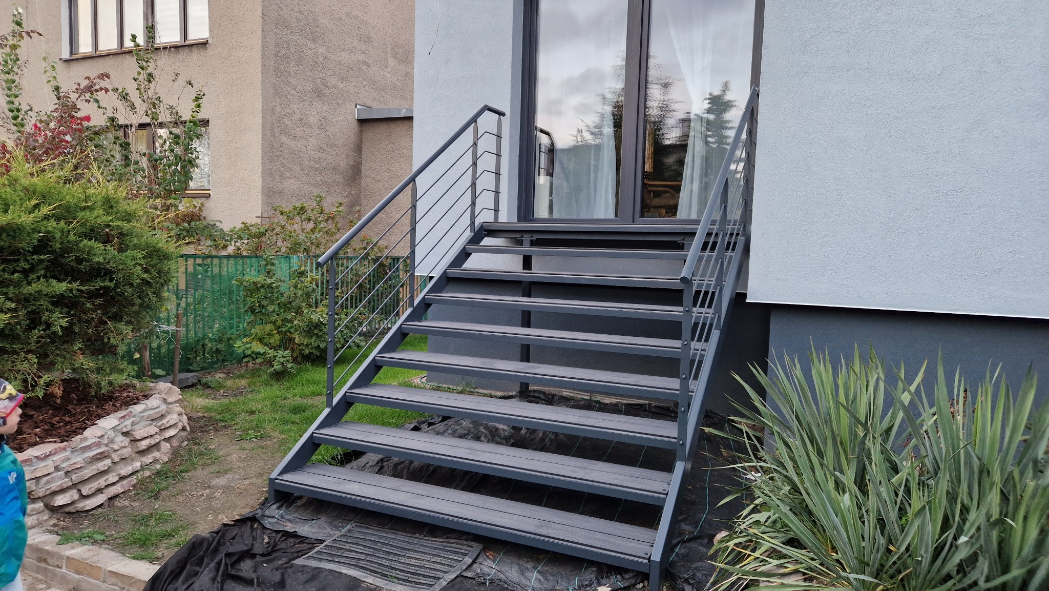 Zewnętrzne schody metalowe z szarą balustradą, prowadzące do drzwi wejściowych budynku. Widoczne rośliny w otoczeniu schodów i fragment elewacji budynku.