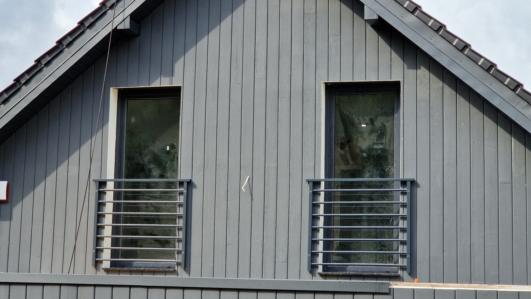 Szara elewacja domu z dwoma oknami zabezpieczonymi poziomymi, metalowymi balustradami. Widok na fragment dachu i niebo.