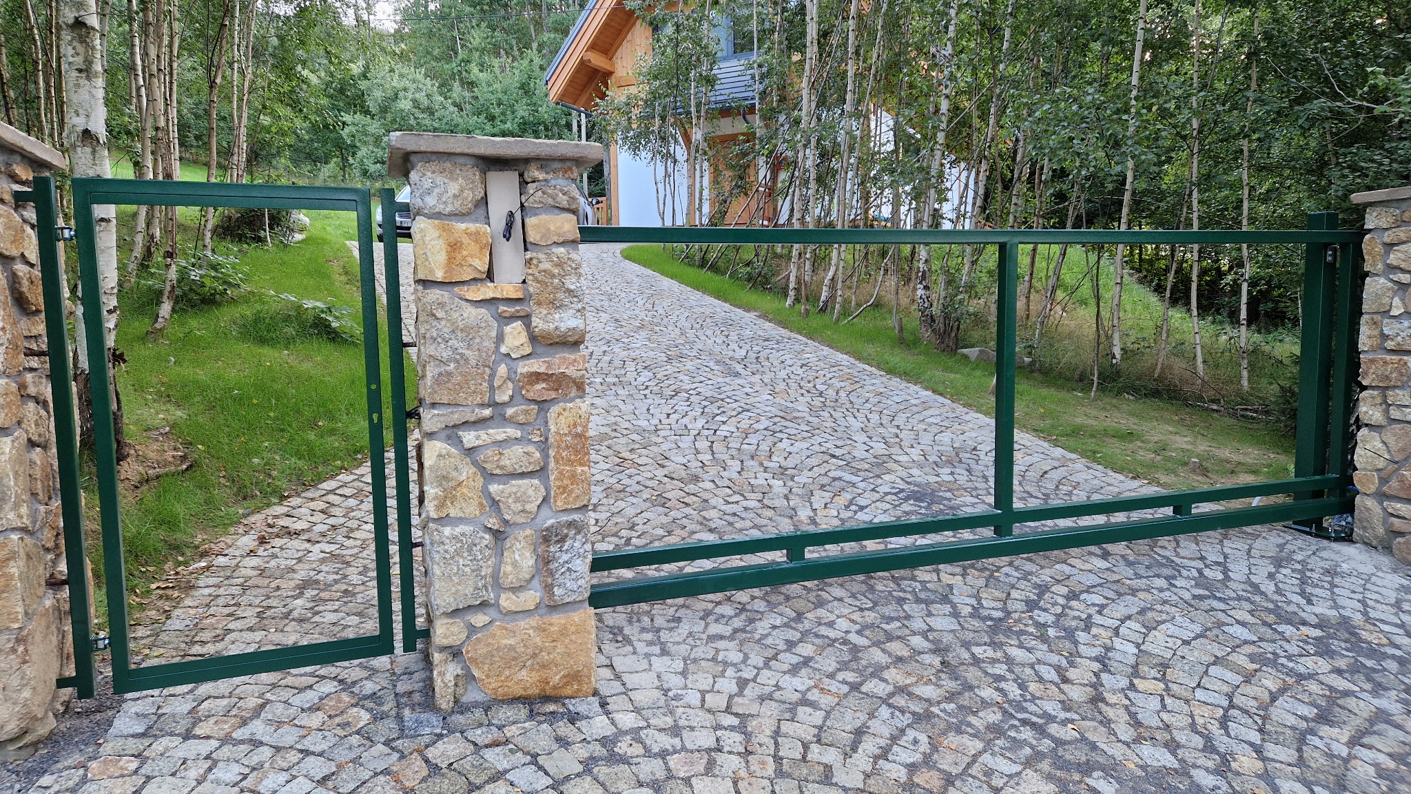 Otwarta, zielona brama wjazdowa z metalu, osadzona na kamiennych słupkach, prowadząca na wybrukowany podjazd w kierunku domu otoczonego zielenią.