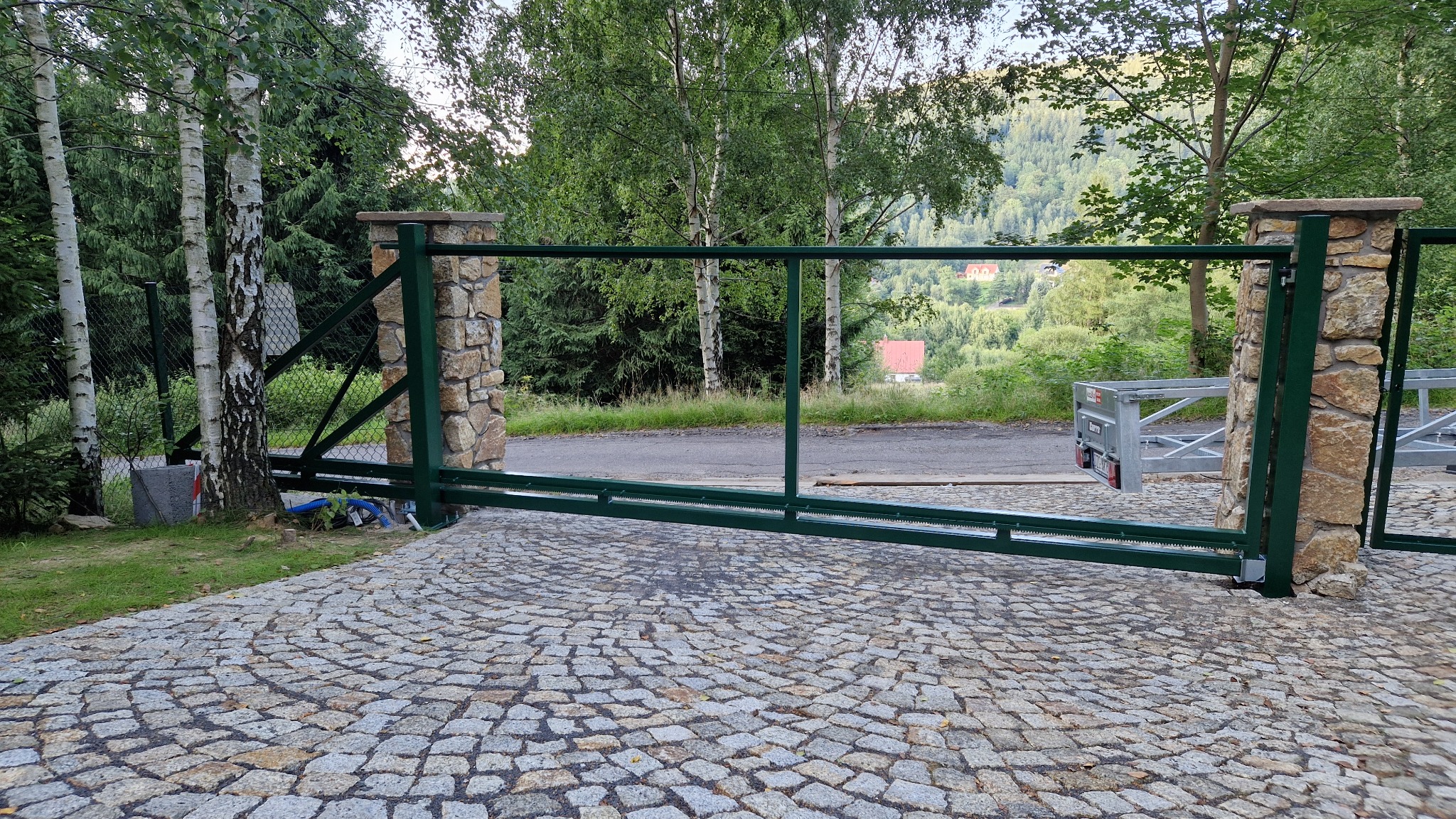 Otwarta, zielona brama wjazdowa z kamiennymi słupkami, prowadząca na posesję z brukowaną nawierzchnią. W tle zieleń drzew i zabudowania.