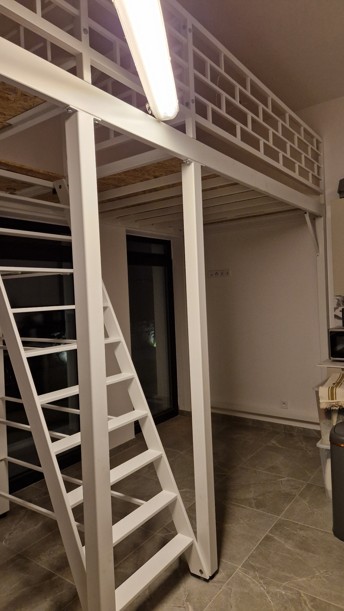 Biała, metalowa konstrukcja antresoli z drabinką w jasnym wnętrzu. Widoczne elementy mocujące i oświetlenie. Minimalistyczny design.