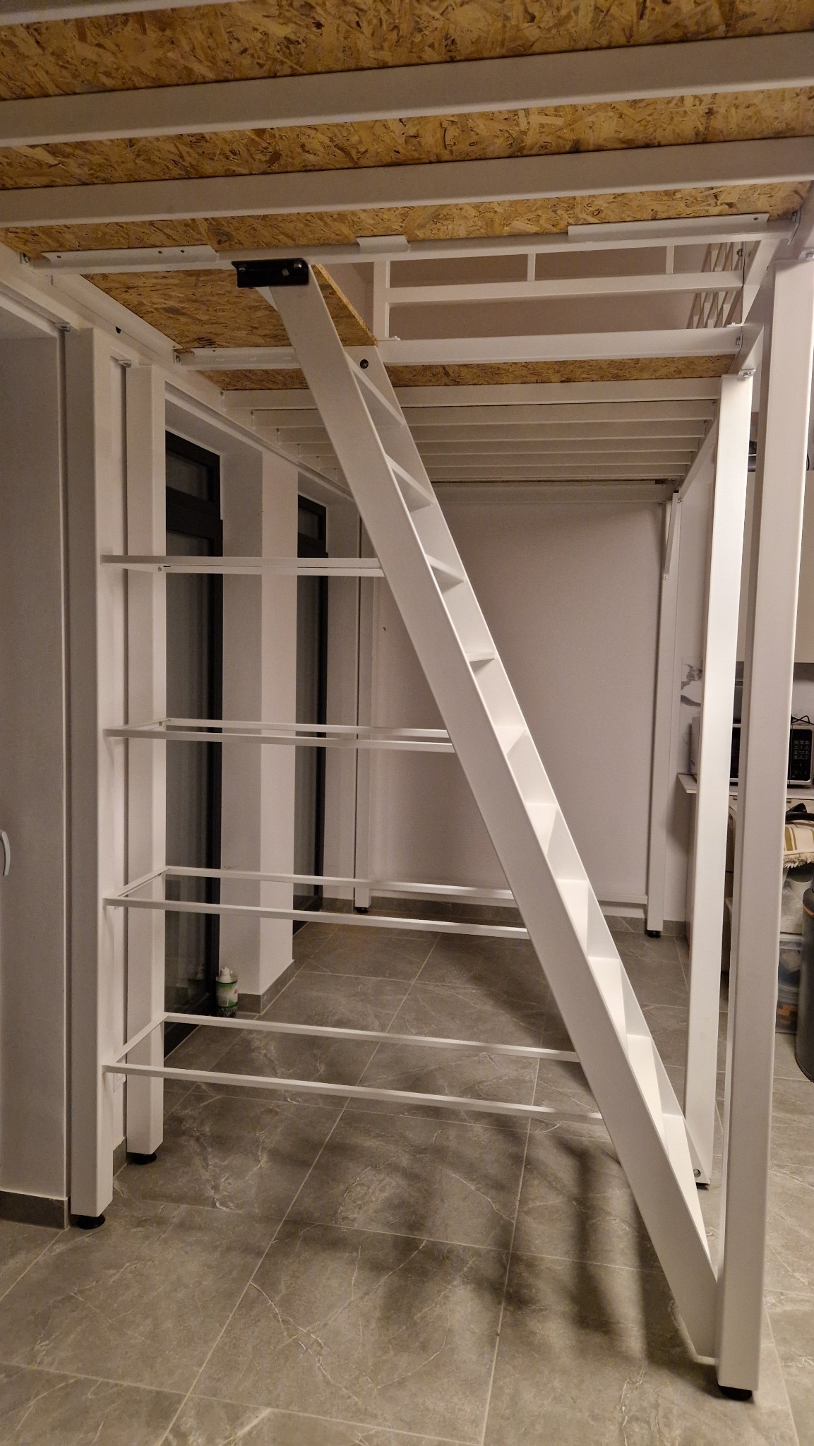 Biała, metalowa konstrukcja antresoli z drabinką i półkami, widok od dołu, na tle szarej podłogi i białych ścian. Widoczna płyta OSB jako sufit antresoli.