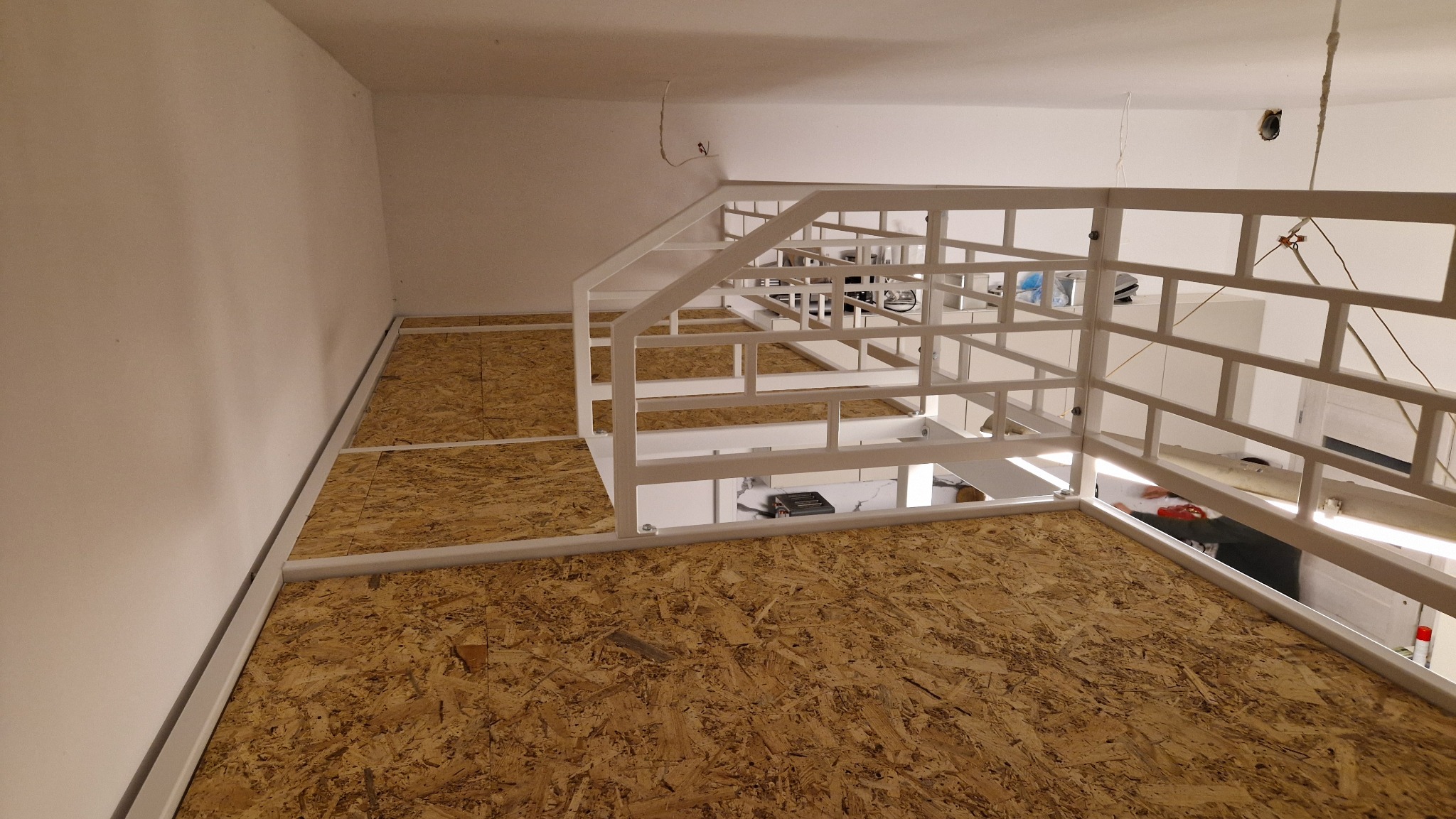 Wnętrze z antresolą: biała, metalowa konstrukcja z drabinką i drewnianą podłogą z płyty OSB. Widok z góry, surowe wykończenie, minimalistyczny design.