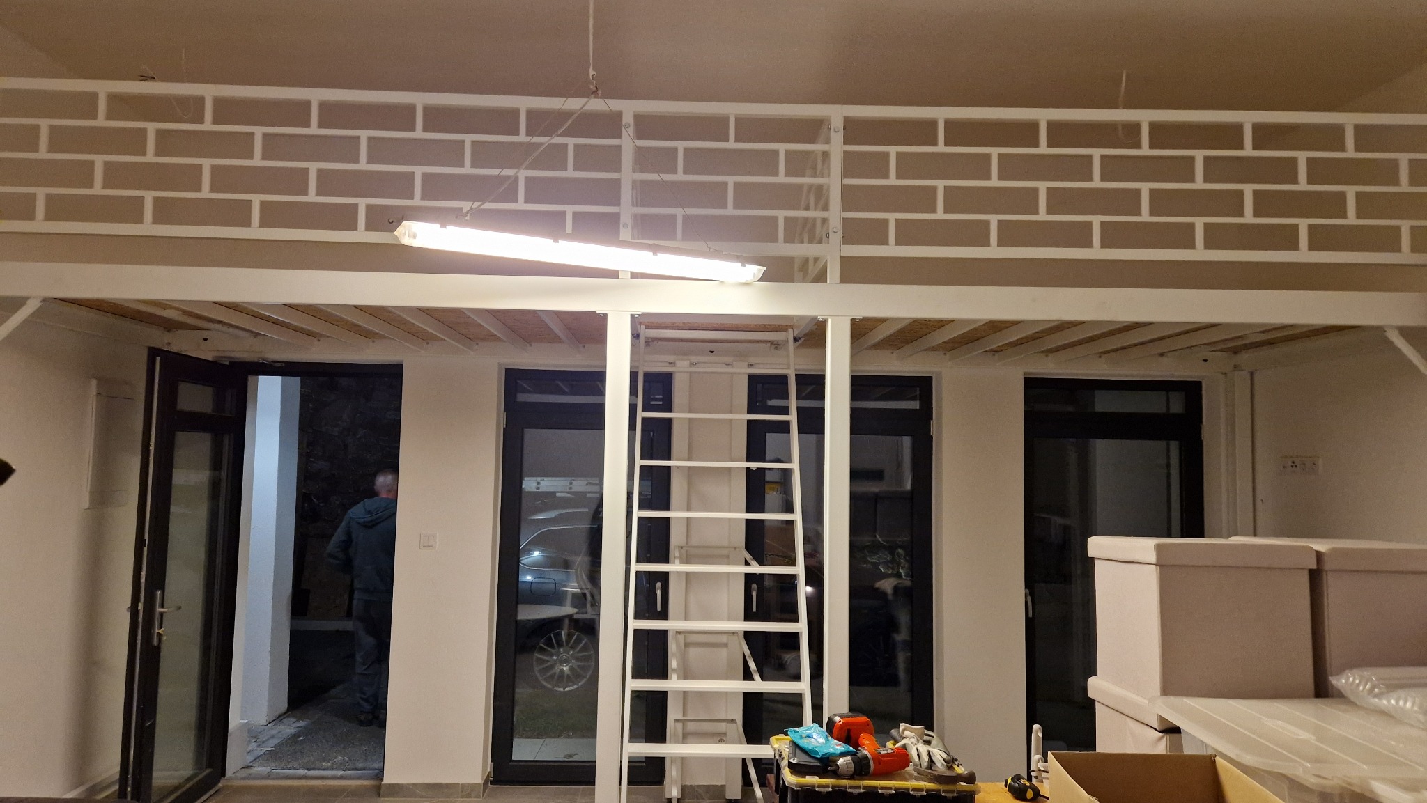 Biała, metalowa konstrukcja antresoli z drabinką w jasnym pomieszczeniu. Widoczne okna i drzwi balkonowe. Na pierwszym planie narzędzia.