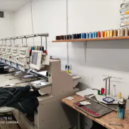 Przemysłowa pracownia z rzędem maszyn do haftowania, na stole leżą czarne bluzy z kapturem, na półkach widoczne szpule nici w różnych kolorach oraz pudełka nici firmy Madeira.