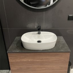 I WOOD DESIGN - Nowoczesna łazienka z umywalką nablatową w kształcie owalu, czarną armaturą i szafką pod umywalką z drewnopodobnym frontem, uzupełniona okrągłym lustrem i szarą glazurą.