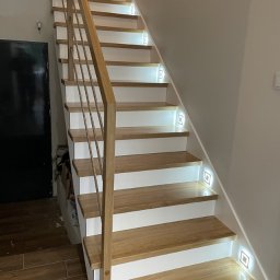 I WOOD DESIGN - Drewniane schody z białymi podstopnicami i podświetleniem LED wbudowanym w ścianę, metalowa poręcz, widoczny fragment podłogi z płytek imitujących drewno.