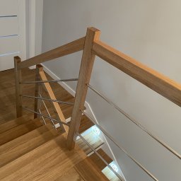 I WOOD DESIGN - Nowoczesne schody z drewnianymi stopniami i balustradą z drewnianych słupków oraz poziomych, metalowych prętów, widok z góry na fragment biegu schodów, z widocznym fragmentem drzwi w tle.