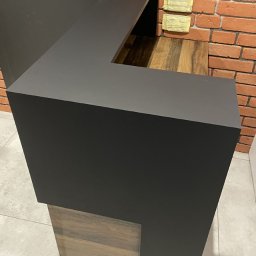 I WOOD DESIGN - Ciemny, matowy blat w kształcie litery L, z drewnianą podstawą, na tle ceglanej ściany i ciemnych drzwi z metalową klamką. Widoczne dwa zabezpieczone gniazdka elektryczne na ścianie oraz logo firmy...