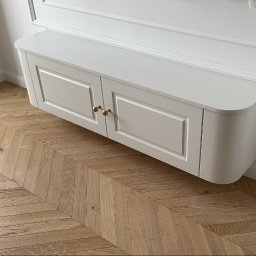 I WOOD DESIGN - Biała, podwieszana szafka z frezowanymi drzwiczkami i złotymi gałkami na tle parkietu ułożonego we wzór jodełki oraz białej ściany z dekoracyjną listwą.
