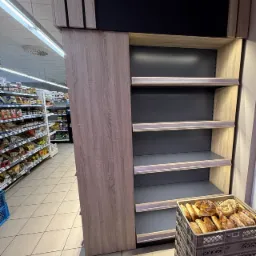 Regał sklepowy z płyty meblowej w supermarkecie w Białymstoku. Półki szare, góra czarna z elementami drewnianymi. W koszyku pieczywo.