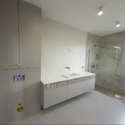 Nowoczesna łazienka z beżowymi szafkami na wymiar, złotą armaturą i kabiną prysznicową z marmurowymi płytkami. Minimalistyczny design z dbałością o detale.