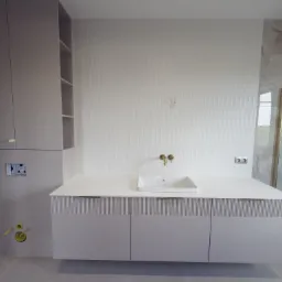 Jasna łazienka z szafką pod umywalką z frezowanymi frontami, złotą armaturą i płytkami imitującymi marmur w strefie prysznica. Nowoczesny design.